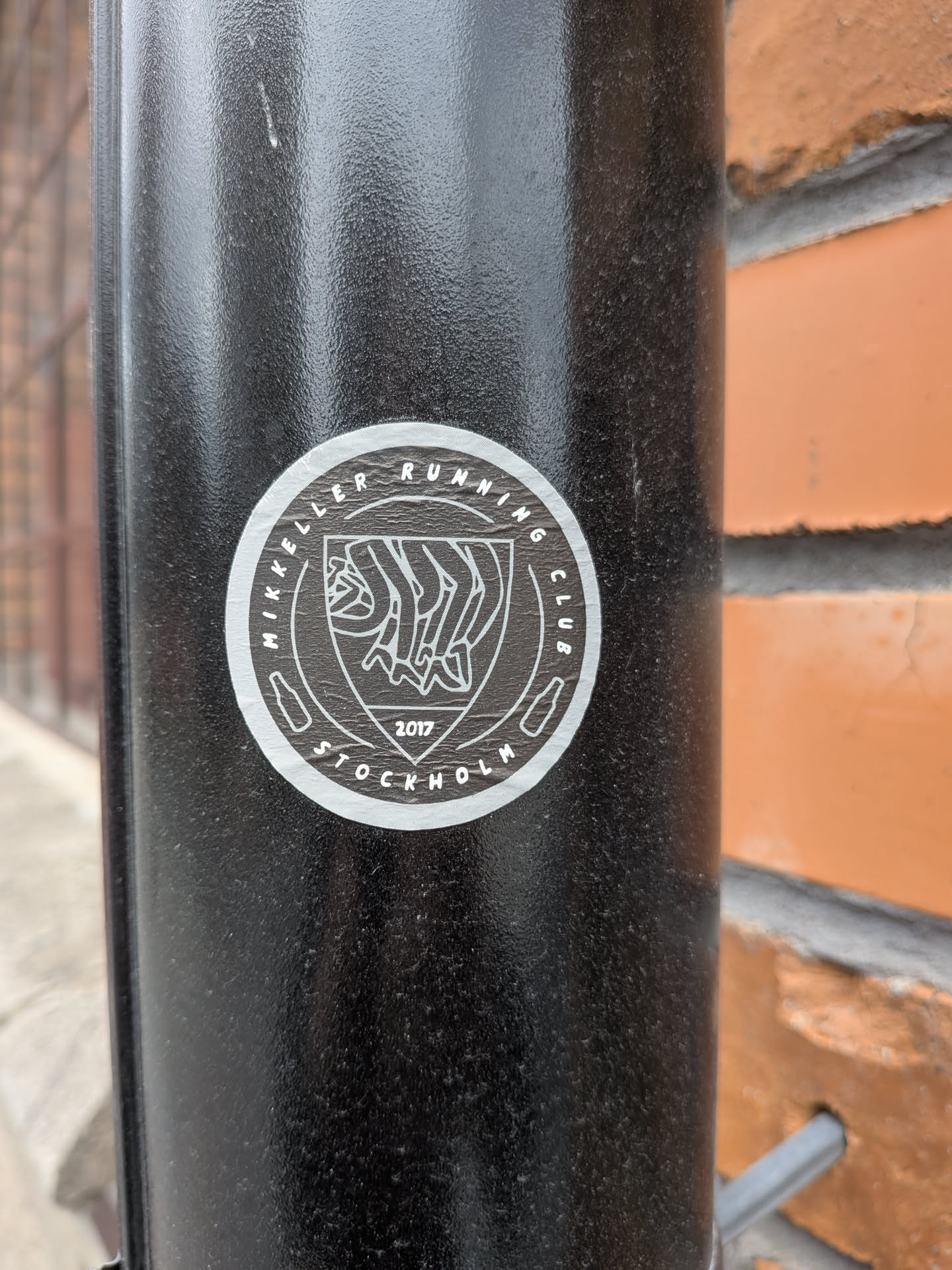 The sticker is circular with a white border and a white outline. The center features a shield-like emblem with a stylized depiction of running legs in white lines. Above the emblem, the text 'MIKKELER RUNNING CLUB' is written in a circular fashion. Below the emblem, the year '2017' is prominently displayed, and further down, 'STOCKHOLM' is also written in a circular manner. Small icons resembling beer bottles are placed on either side of the emblem within the circular text.