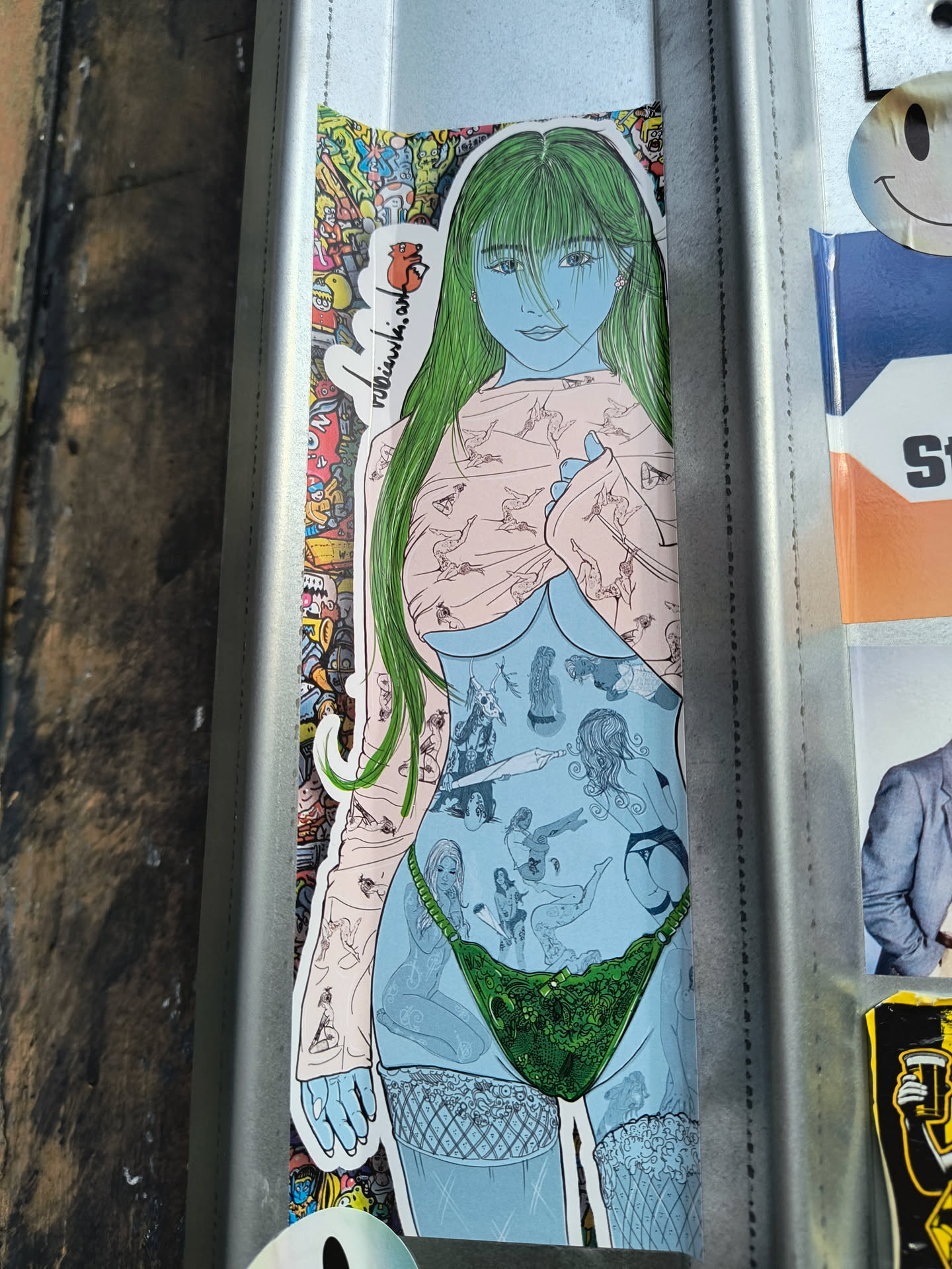 A sticker featuring a stylized illustration of a woman with long green hair. Her body is adorned with various smaller illustrations.  The style is reminiscent of graphic novels or anime. The sticker is affixed to a metal surface alongside other stickers.