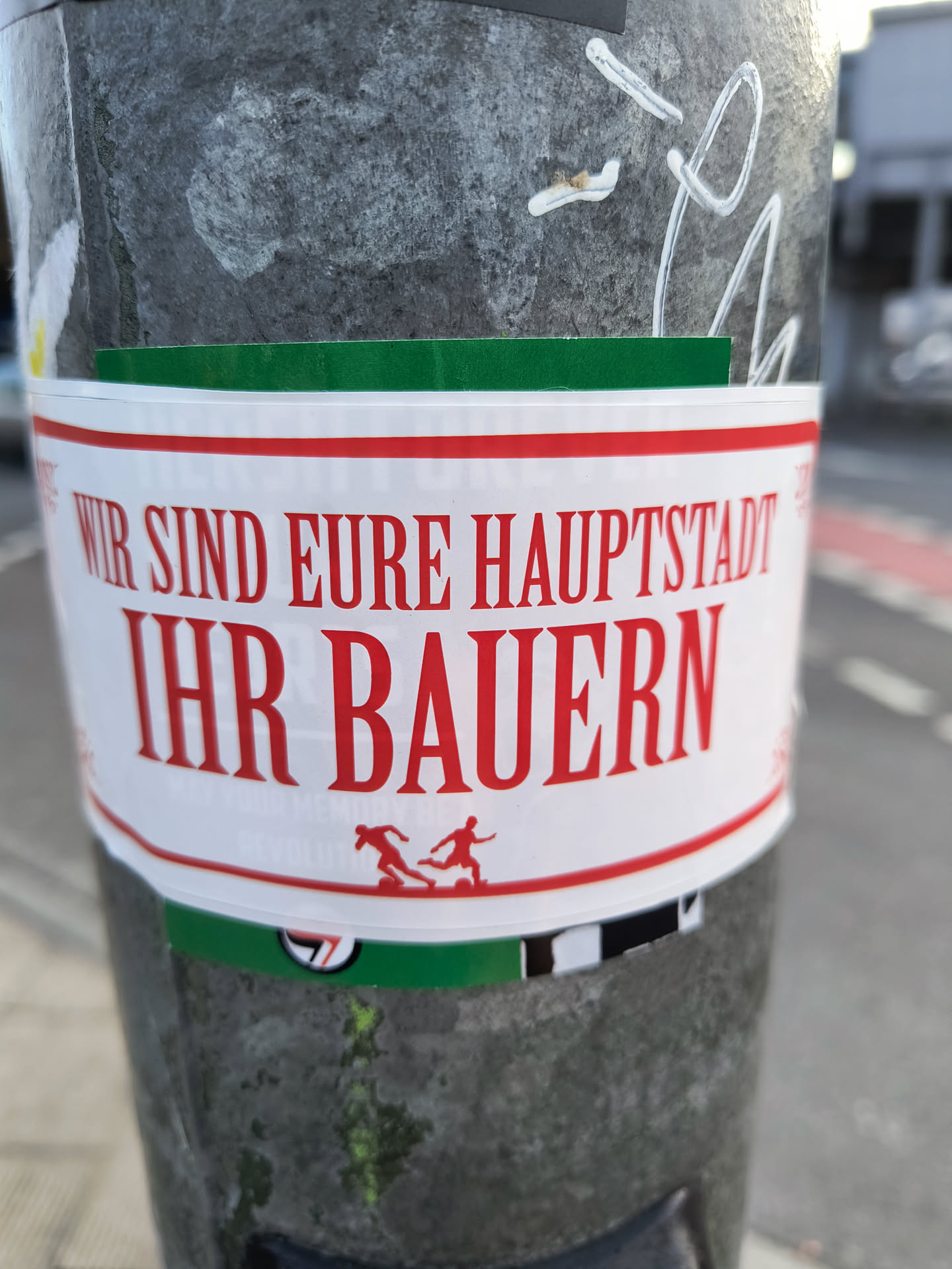 A sticker on a pole with a bold text in German that translates to "We are your capital, your farmers." It also features a small graphic of two figures seemingly running or playing. The sticker has a green stripe at the top and bottom.