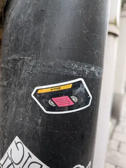 The sticker is shaped like a VHS cassette tape. It features a yellow label area at the top with black text. Below this, there is a black section representing the tape housing, with two circular spools visible. A bright pink rectangular area with horizontal black lines is positioned in the center, simulating the magnetic tape. The sticker has a white border and appears to be applied to a dark, textured surface.