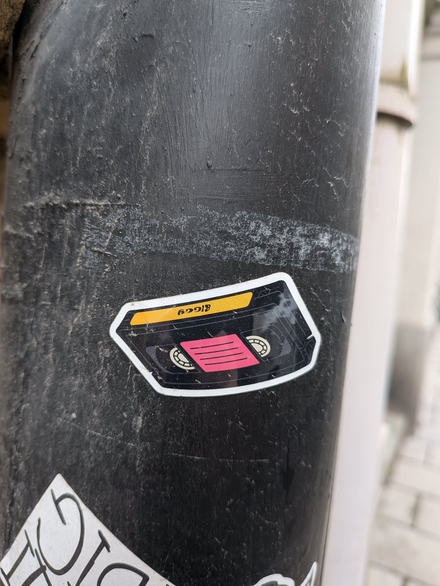 The sticker is shaped like a VHS cassette tape. It features a yellow label area at the top with black text. Below this, there is a black section representing the tape housing, with two circular spools visible. A bright pink rectangular area with horizontal black lines is positioned in the center, simulating the magnetic tape. The sticker has a white border and appears to be applied to a dark, textured surface.
