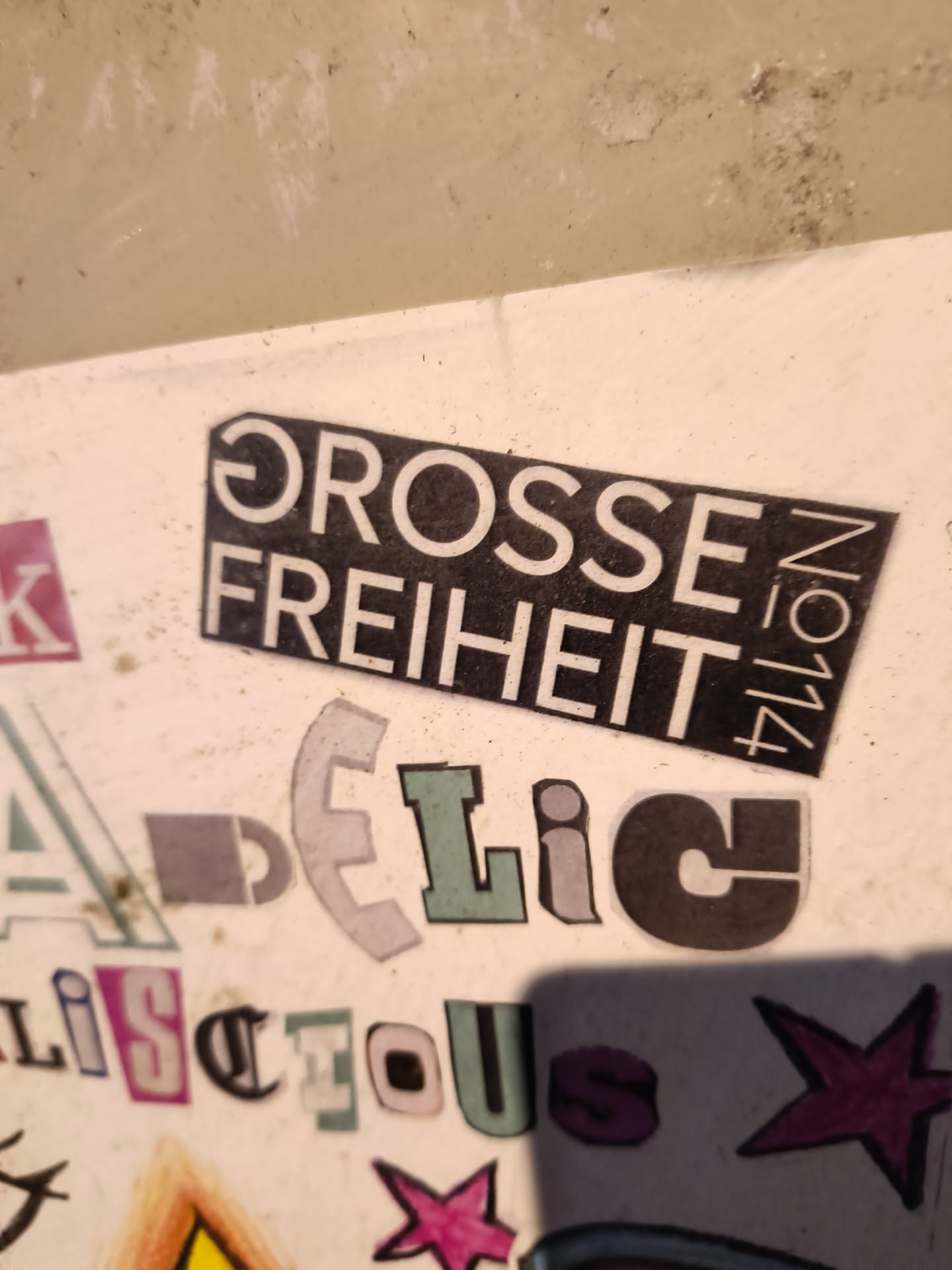 A black rectangle sticker with the words "GROSSE FREIHEIT No. 114" in white, pasted among other stickers on a white surface. The other stickers are various letters cut out from magazines, giving a collage effect.