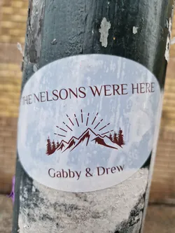 Oval white sticker with brown text and graphic.  It says "The Nelsons Were Here" at the top, features a mountain range with sunburst graphic in the center, and "Gabby & Drew" at the bottom.