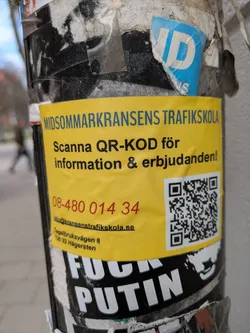 A yellow sticker on a pole advertises Midommarkransens Trafikskola driving school. It urges people to scan the QR code for information and offers, and provides a phone number and email address.