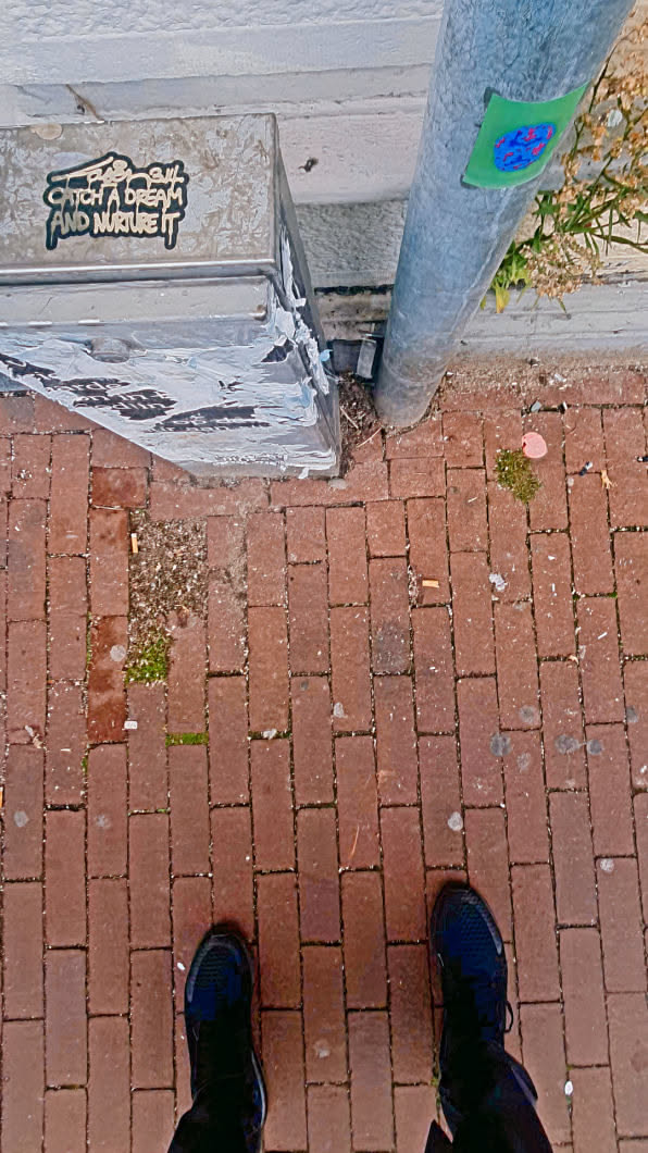 A photo from a top-down angle of a brick road with a metal pipe, a metal electrical box and the shoes of the person taking the photo in the bottom. The electrical box has a sticker that says "Catch a dream and nurture it", and the metal pipe has another sticker of an abstract design.