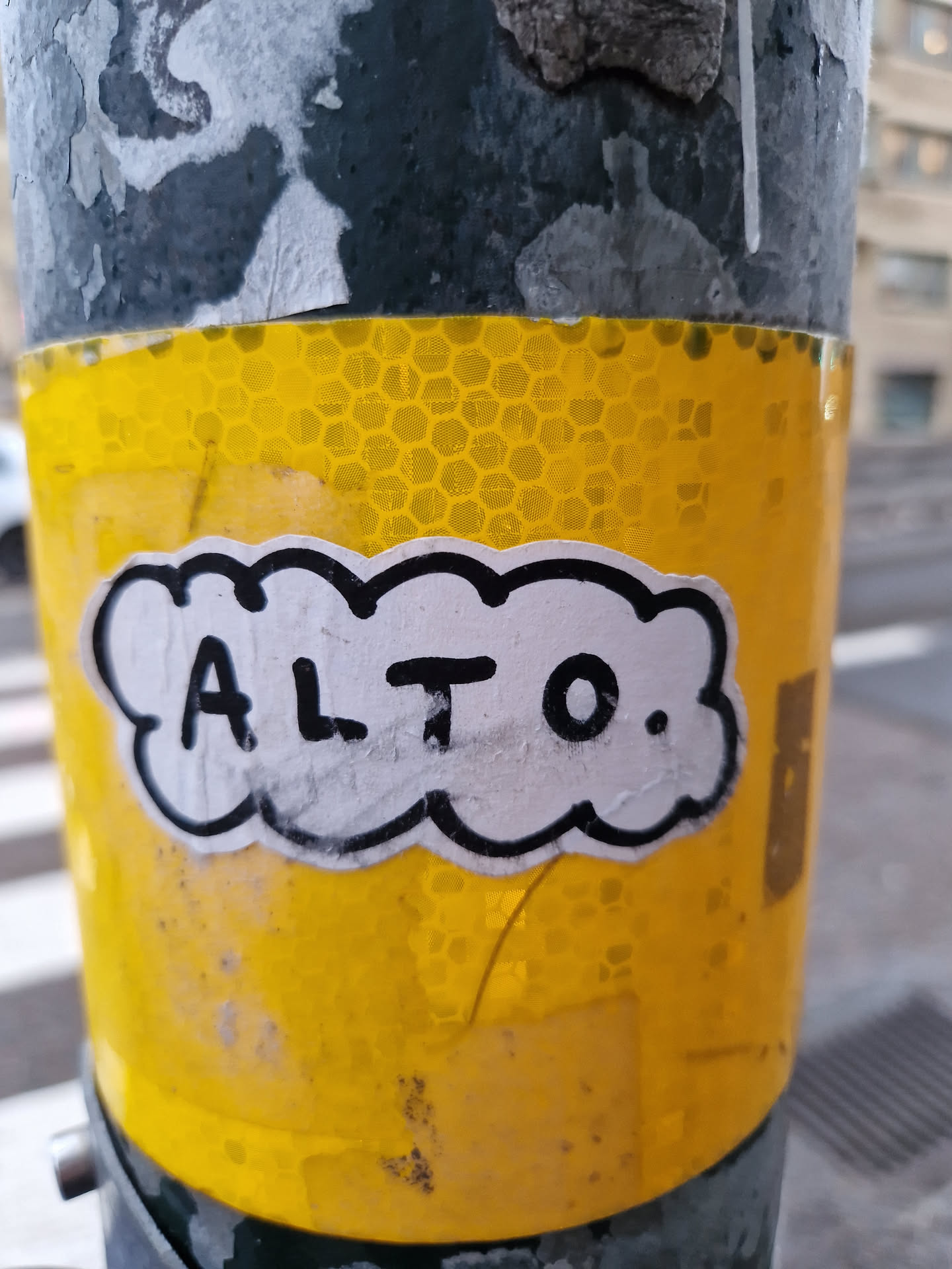 A simple black and white illustration sticker on a yellow reflective surface, with the word "ALTO." written in capital letters.  The style is simple and bold. The sticker is affixed to a pole, suggesting it's a street art piece.