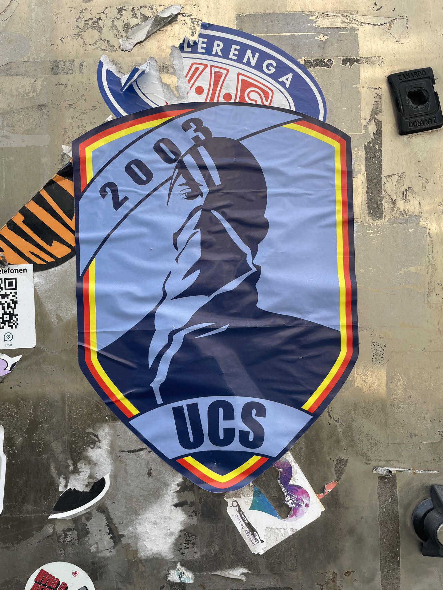 A shield-shaped sticker featuring a dark blue silhouette of a hooded figure, facing right, against a light blue background. The figure's face is mostly obscured, with only a single eye visible. The shield has a prominent border with red and yellow stripes, reminiscent of a flag. The year '2003' is written in dark blue at the top left, following the curve of the shield, and the letters 'UCS' are in dark blue at the bottom center. The sticker shows signs of wear, including creases and wrinkles, indicating it is adhered to a textured wall.