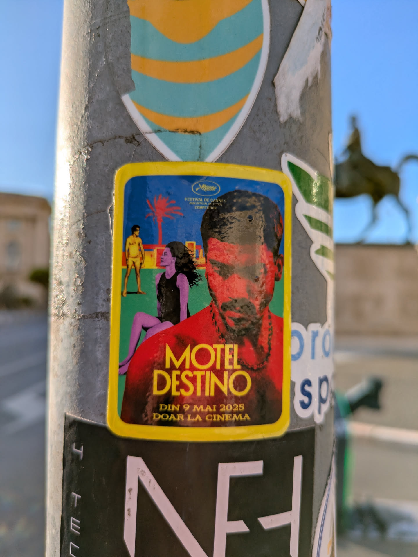 A yellow-bordered sticker on a pole shows a movie poster featuring two figures in swimwear, a woman in a black dress, and a man's face rendered in red tones. The poster reads "Motel Destino" and "Din 9 mai 2025 doar la cinema", also features the Cannes Film Festival logo.