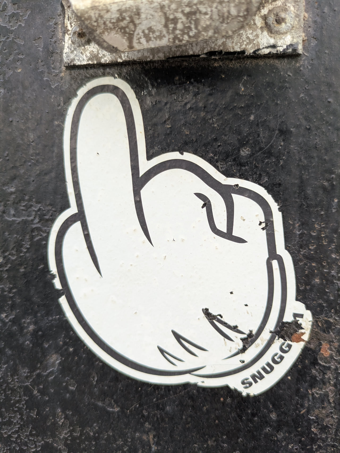 This sticker features a white cartoon hand in a distinctive gesture, likely a play on a common rude hand sign, with black outlines. The word "SNUGG" is written in bold black letters at the bottom right corner of the sticker. The sticker appears slightly worn with some small damages.