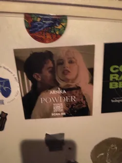 The sticker features a close-up portrait of a man leaning in to whisper to a woman. The man has dark hair and is looking away from the camera. The woman has light-colored hair and is looking directly at the camera with a neutral expression. The background is blurred but suggests an indoor setting. Prominently displayed on the sticker are the words "ARNIKA POWDER" in a modern, sans-serif font. Below the text, a QR code is visible with the words "SCAN ME" below it.