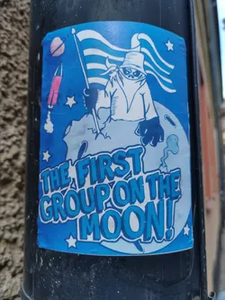A blue sticker featuring a cartoon astronaut figure on the moon, holding a flag.  The style is reminiscent of vintage space exploration imagery. The text 'THE FIRST GROUP ON THE MOON!' is prominently displayed.