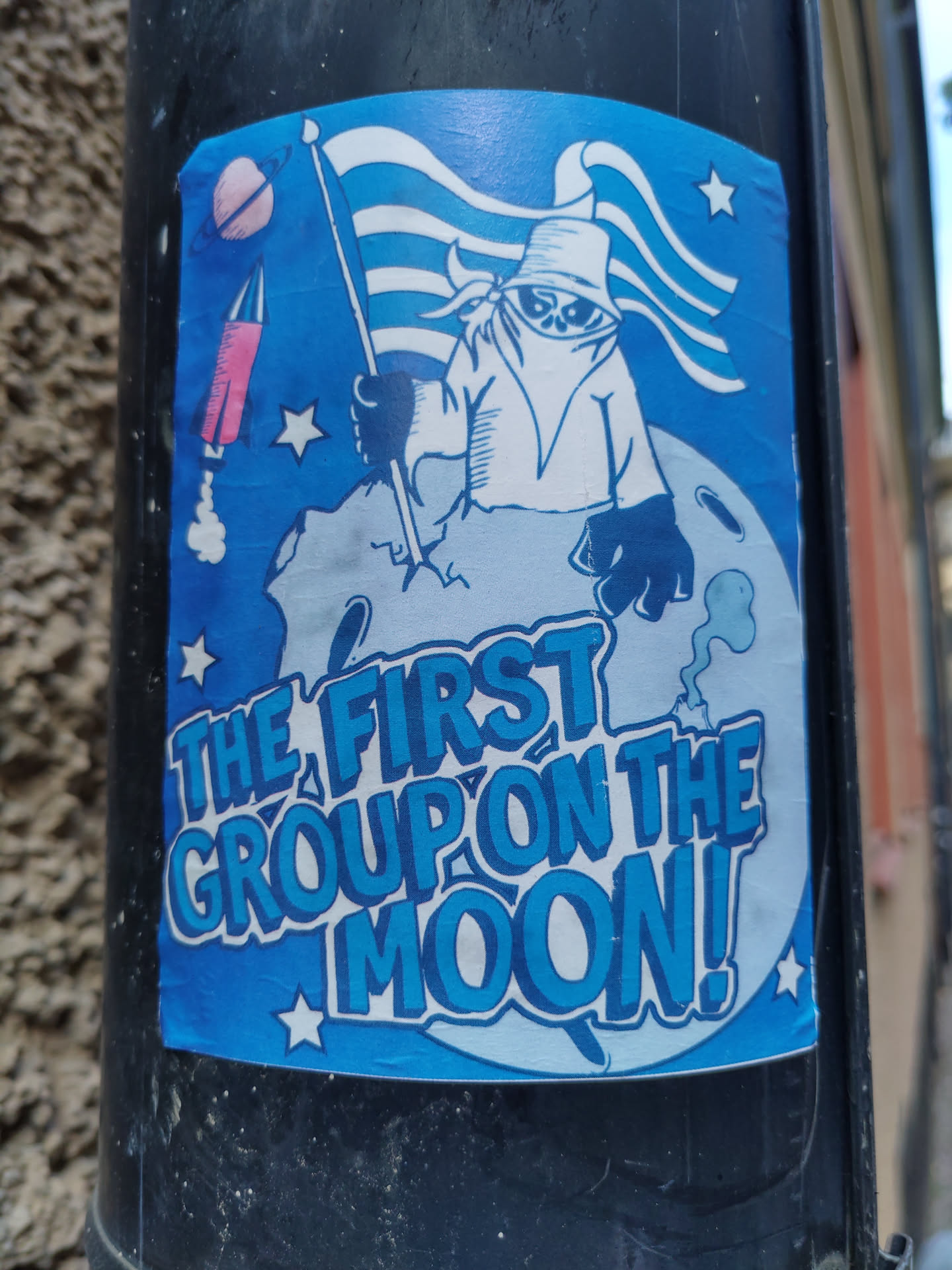 A blue sticker featuring a cartoon astronaut figure on the moon, holding a flag.  The style is reminiscent of vintage space exploration imagery. The text 'THE FIRST GROUP ON THE MOON!' is prominently displayed.