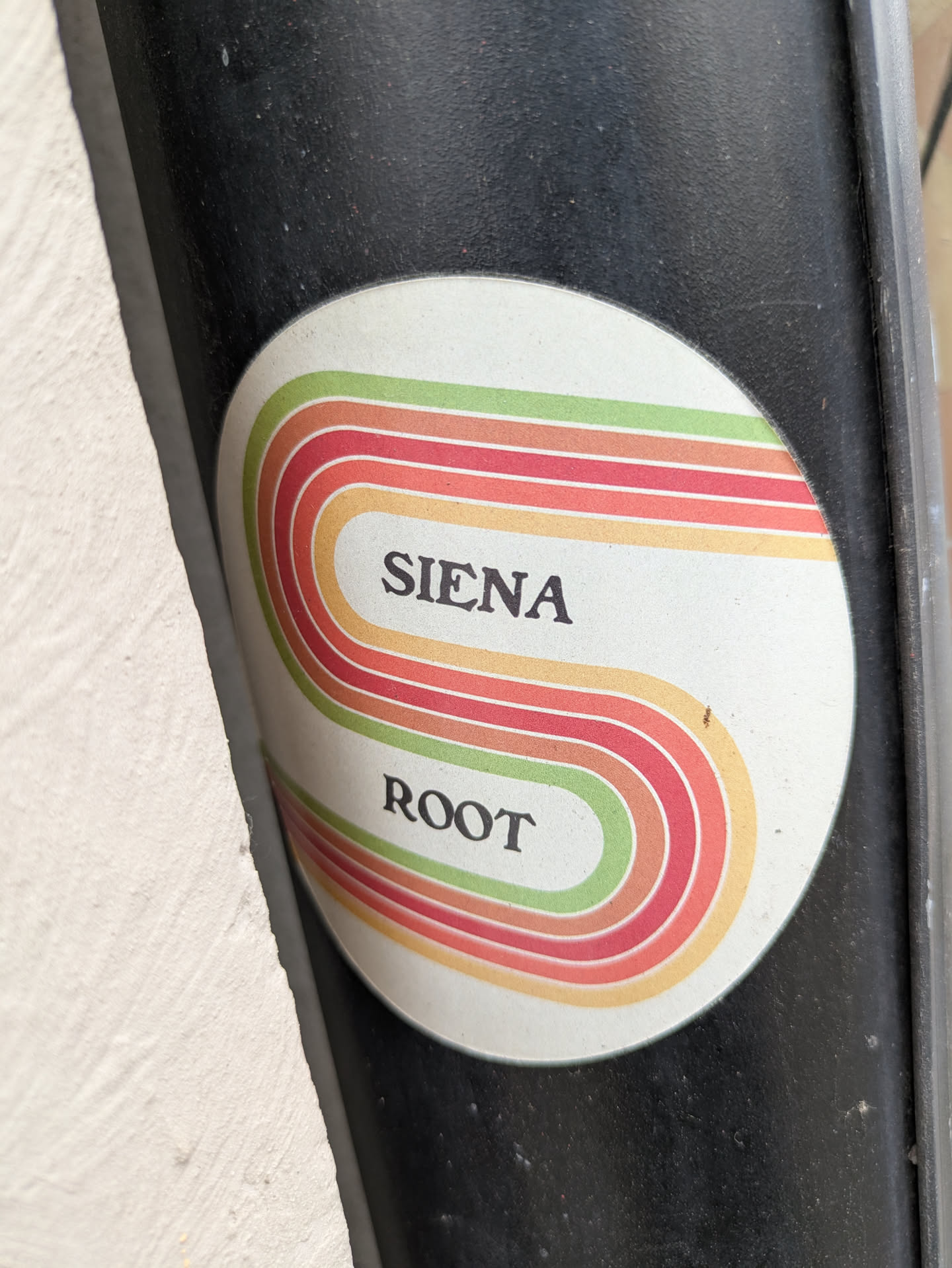 A round sticker with a retro-style design. The sticker features multiple colored stripes, with the colors in the order: green, orange, red, orange forming an S shape. The text "SIENA" and "ROOT" are written in black in between the stripes.