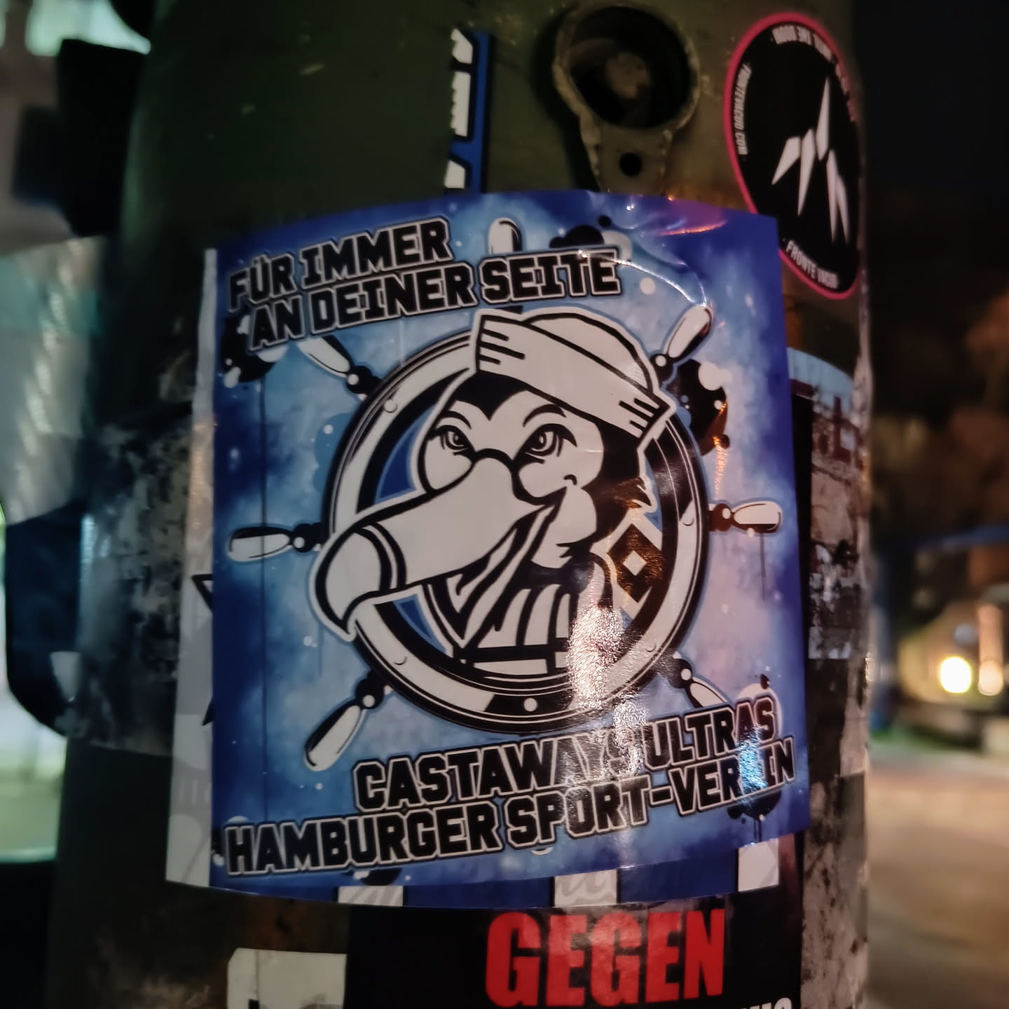 A blue sticker with a cartoon toucan wearing a sailor's hat in the center of a ship's wheel. The text on the sticker says "Für immer an deiner Seite" (Forever by your side) and "Castaways Ultras Hamburger Sport-Verein".