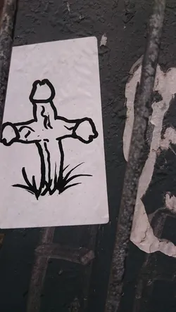 This sticker features a minimalist black and white drawing of a cross with a phallic symbol at its center.  The cross is planted in grass. The style is simple and slightly irreverent.