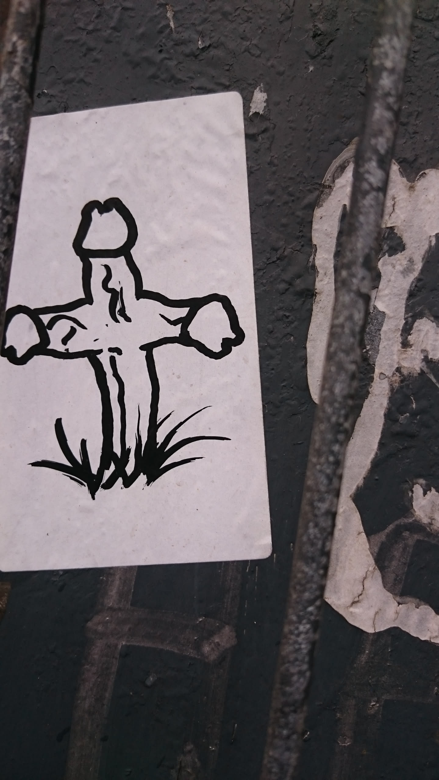 This sticker features a minimalist black and white drawing of a cross with a phallic symbol at its center.  The cross is planted in grass. The style is simple and slightly irreverent.