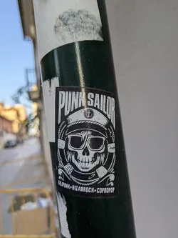 A black and white sticker on a pole features a skull wearing a sailor hat and sunglasses. The text 'Punk Sailor' is at the top, and 'Gilipunk Bizarrock Copropop' is at the bottom. The skull is designed in a punk style.