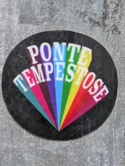 A round sticker on a grey surface. The sticker has a black background with a rainbow-colored design. The words "PONTE TEMPESTOSE" are written in white uppercase letters, arranged in a way that creates a fan-like shape. Each letter is set on a different color of the rainbow.
