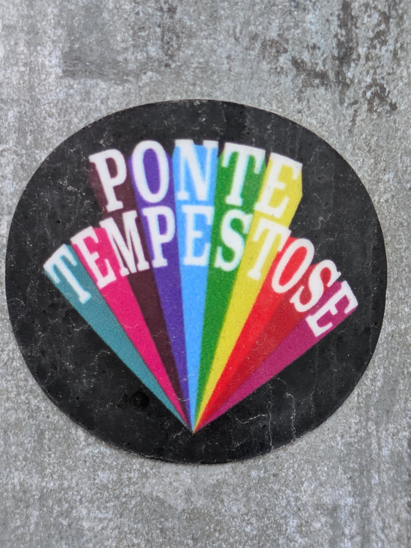 A round sticker on a grey surface. The sticker has a black background with a rainbow-colored design. The words "PONTE TEMPESTOSE" are written in white uppercase letters, arranged in a way that creates a fan-like shape. Each letter is set on a different color of the rainbow.