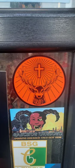 A circular sticker featuring the iconic Jägermeister logo. The background is a vibrant, solid orange, from which dark, radiating lines emanate from behind a black cross positioned centrally between the antlers of a majestic stag's head. The stag's head, including its detailed antlers, ears, and facial features, is rendered in a dark, solid color, likely black or deep brown, with clear outlines. The overall design is bold and instantly recognizable.