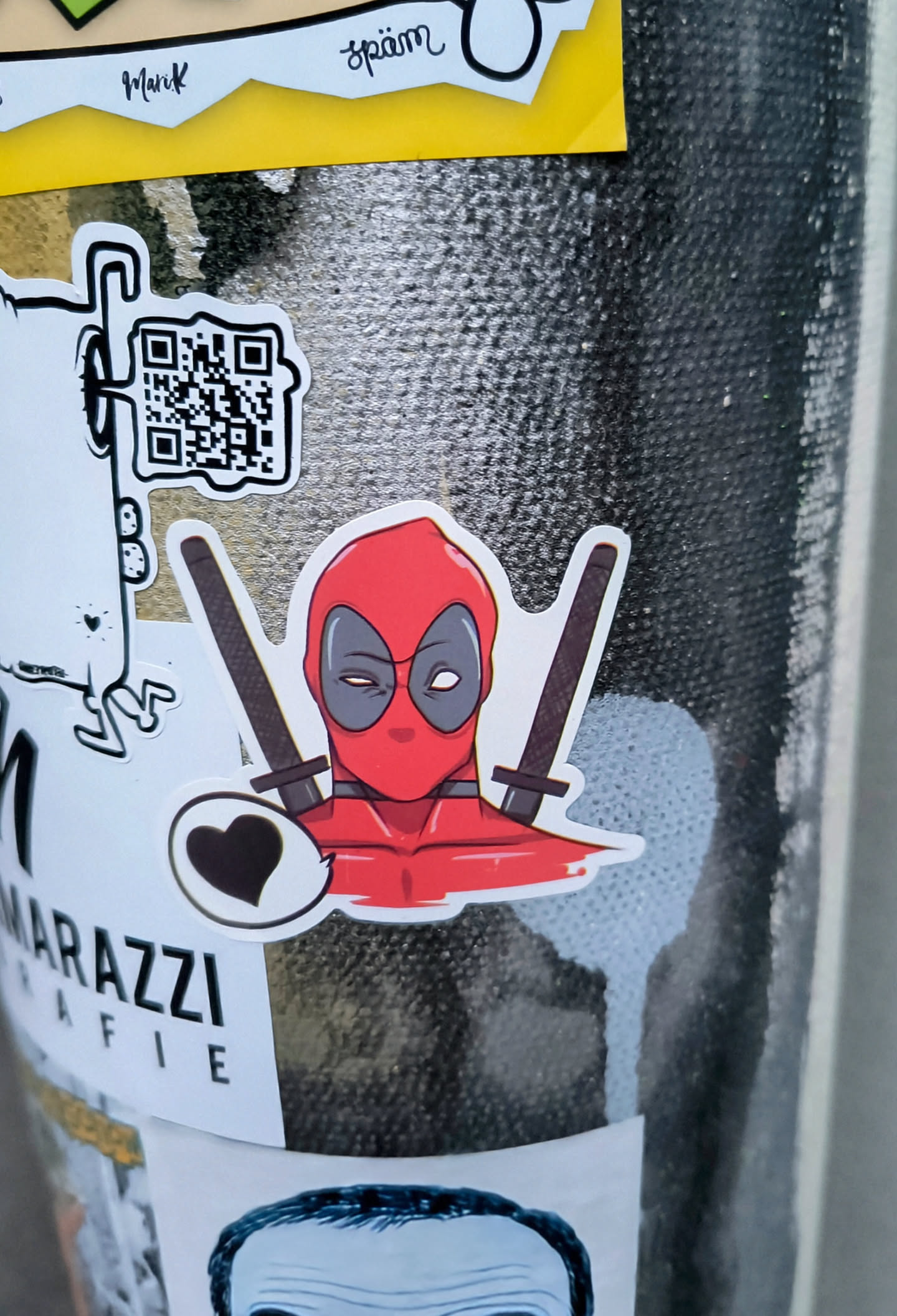 A sticker featuring Deadpool winking, with his katanas behind him and a heart in a speech bubble.  A QR code is visible on another sticker nearby.