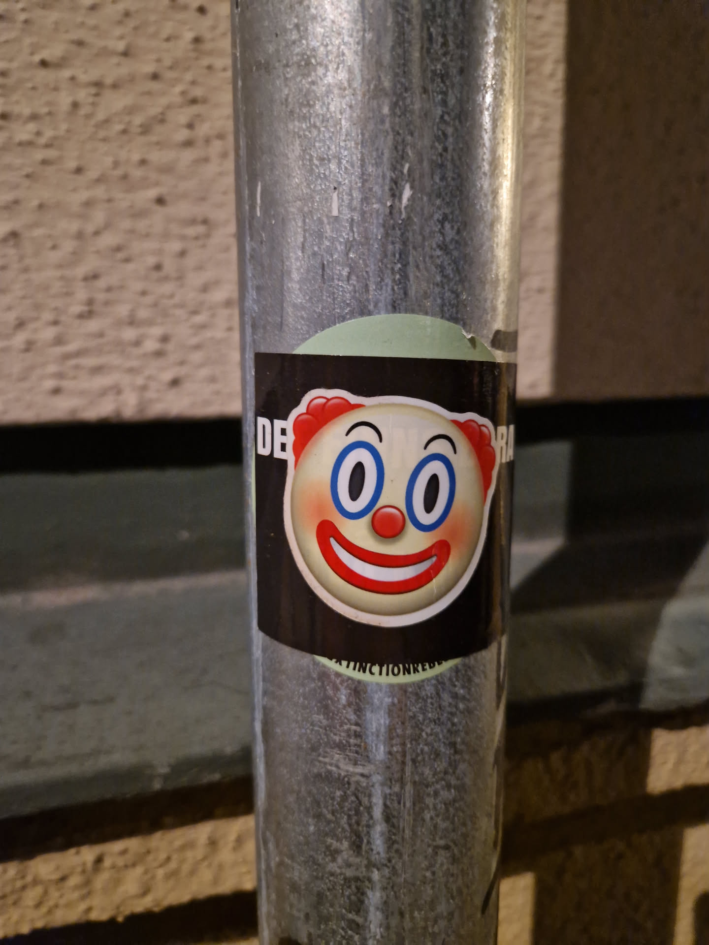 A round sticker featuring a cartoon clown emoji with big blue eyes and a red smile. The sticker is on a dark background with a light green circle behind it. Some text is partially visible on the edges of the sticker.