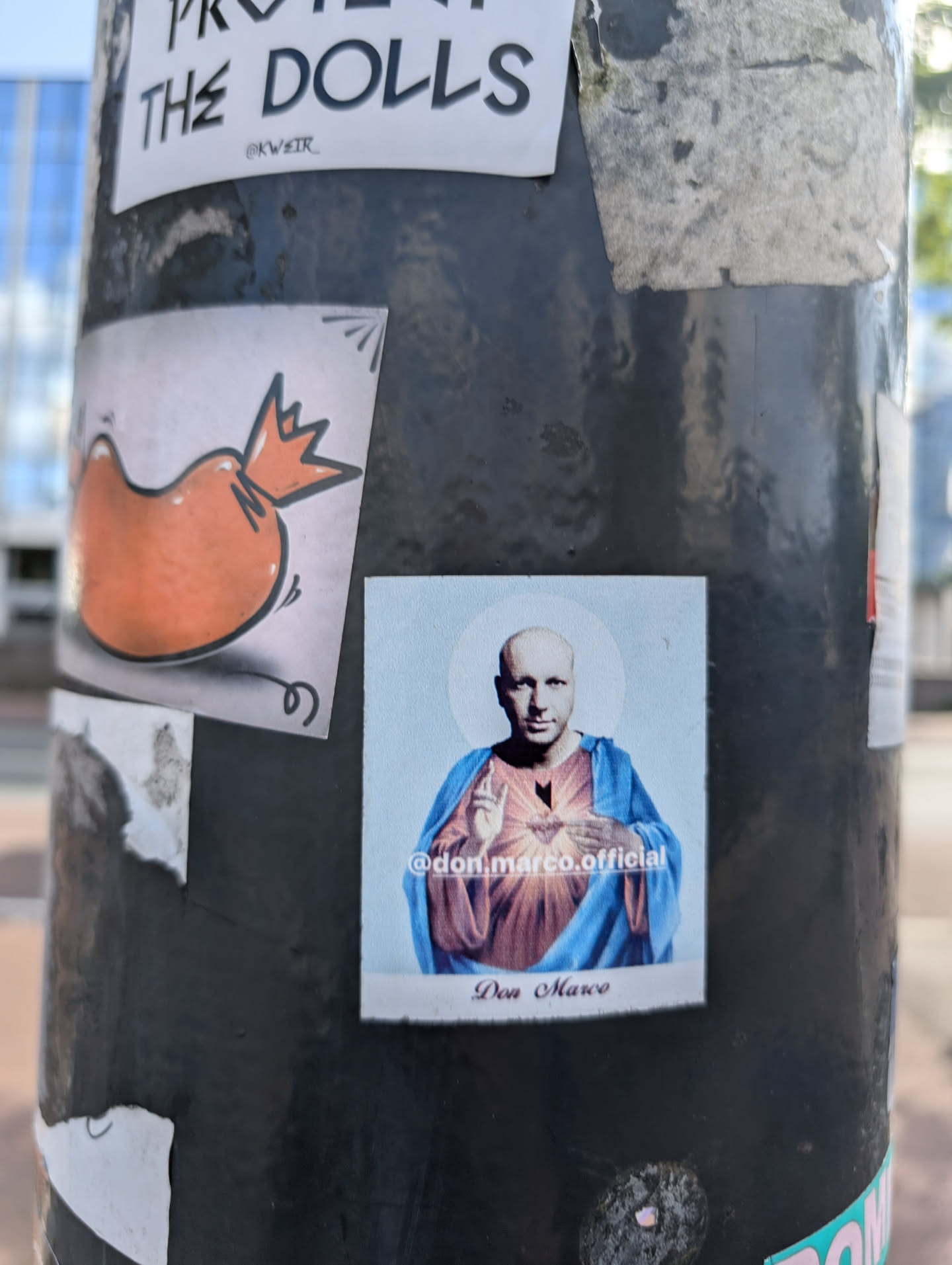 A sticker on a pole features a man with a shaved head, dressed in a blue and red robe, with a halo above his head, resembling Jesus. The sticker has a white border and is labeled with the text "Don Marco" at the bottom, and the instagram handle "@don.marco.official" above the name.
