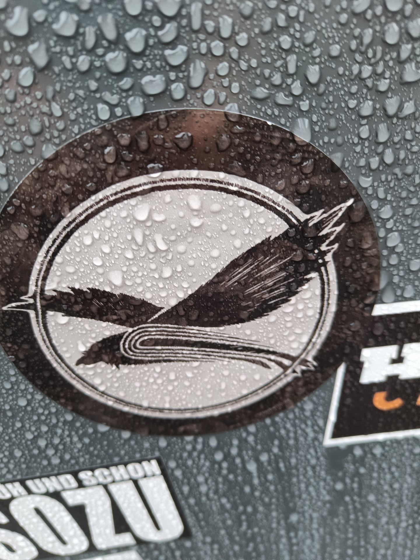 A round black and white sticker featuring a stylized bird design. The bird is depicted in flight with its wings outstretched, designed in a minimalist style. The sticker is wet, with water droplets visible on its surface.