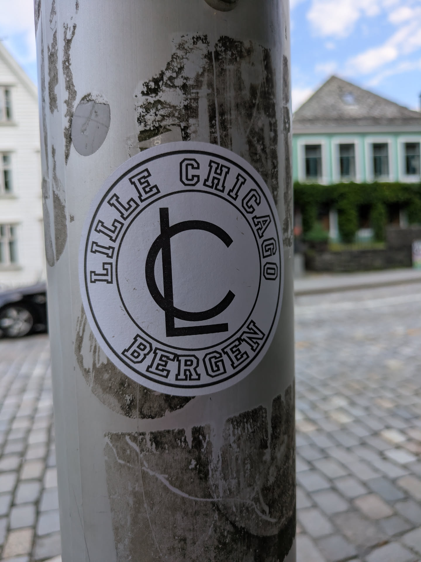 The sticker is round with a white background and thick black outlines. The words 'LILLE CHICAGO' are written in a curved arc along the top edge, and 'BERGEN' is similarly curved along the bottom edge, both in a bold, blocky sans-serif font. In the middle of the sticker, there is a prominent black stylized logo, appearing as an 'L' integrated into a 'C'. The sticker is adhered to a light-colored, possibly metallic pole, which shows signs of wear and tear, including some peeling and discoloration from previous adhesives or dirt. The background is slightly blurred, revealing parts of buildings and a cobbled street under a bright sky.