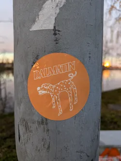 An orange round sticker with a cartoon drawing of a Dalmatian and the word Dalmatin written on it. The sticker is on a grey pole outdoors.