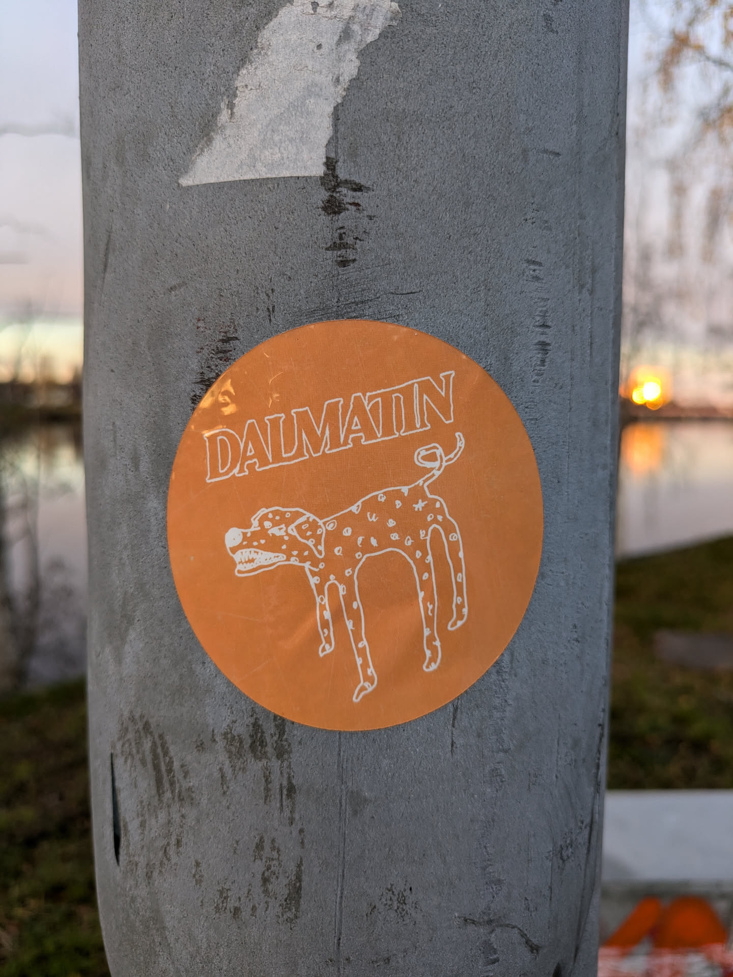 An orange round sticker with a cartoon drawing of a Dalmatian and the word Dalmatin written on it. The sticker is on a grey pole outdoors.