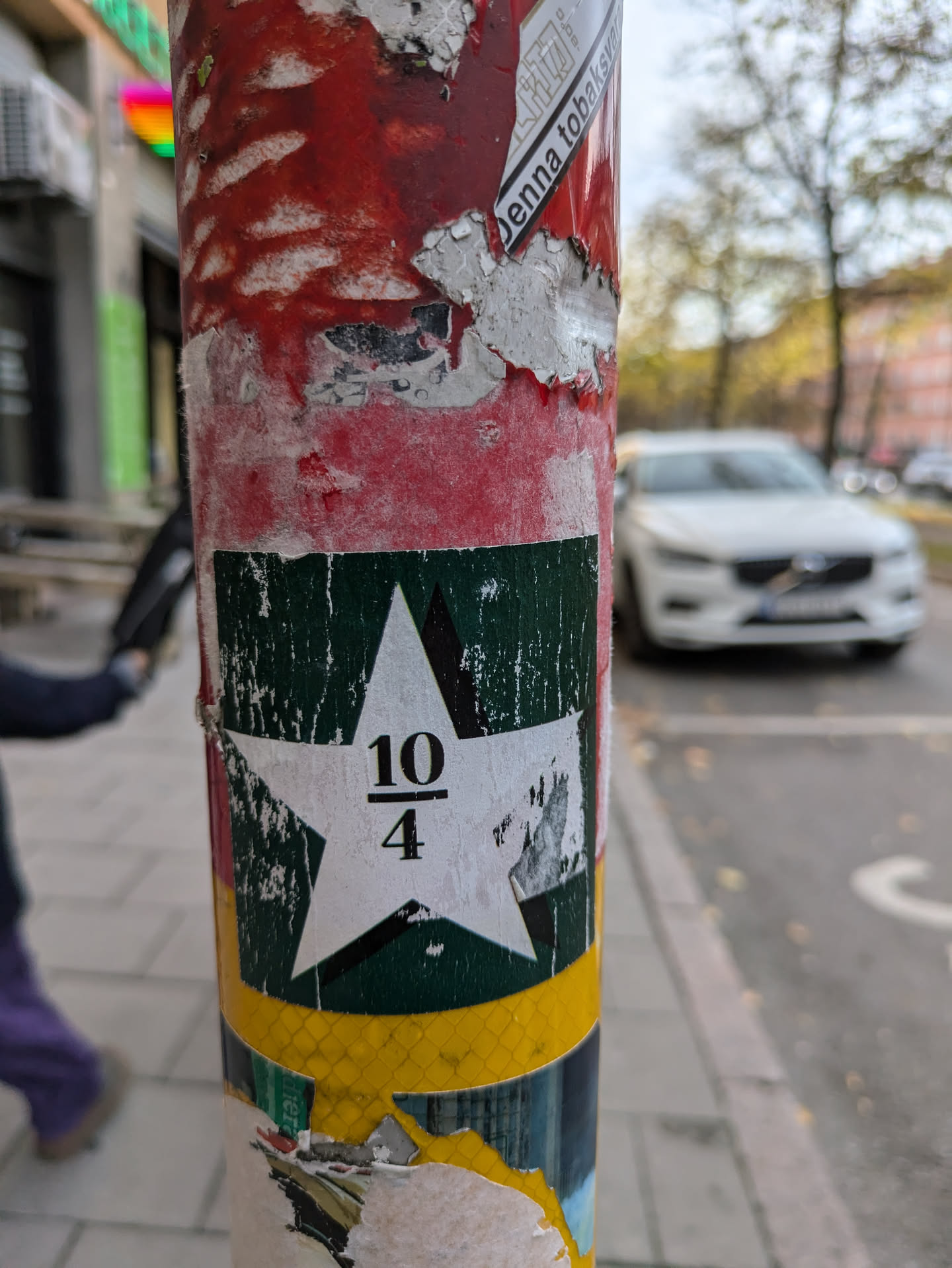 A square sticker with a white star on a dark green background. The fraction 10/4 is printed in the center of the star. The sticker is affixed to a pole covered with other stickers and debris.