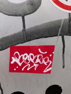 This image shows a red sticker with white stylized writing. The sticker is rectangular and affixed to a surface that appears to be painted in metallic silver. The white writing is somewhat abstract and cursive, with connected letters forming a unique signature or tag. There are also a few small dots around the writing. The sticker is positioned amidst other street art elements. The background is blurred and has a slightly industrial look due to the silver paint and the presence of dark shapes and drips. Other stickers are also visible in the blurred background.