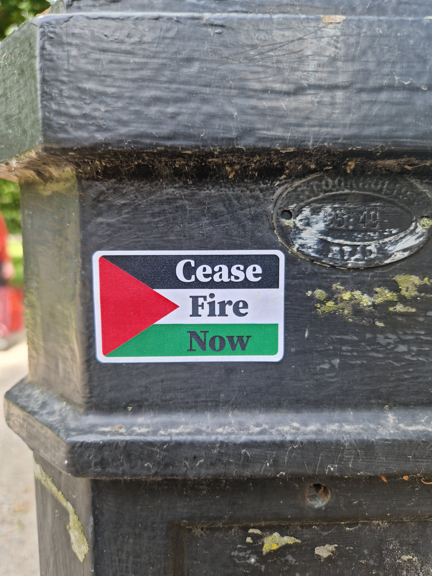 A sticker with a flag-like design and the text "Cease Fire Now". The flag features red, black, and green colors, possibly representing a specific cause or movement related to peace and conflict resolution.