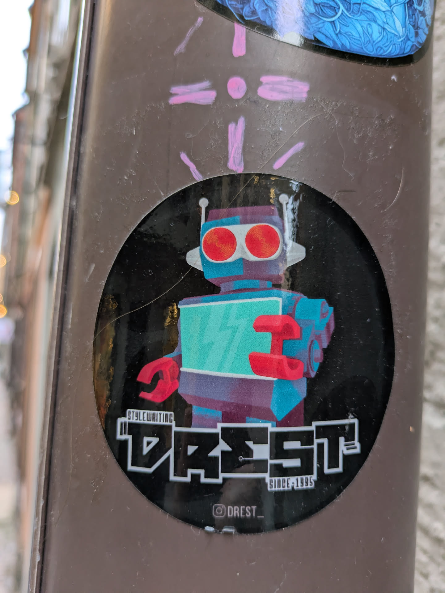 Round sticker featuring a retro-style robot illustration with a teal body, red arms, and large red eyes. The text "DREST." is prominently displayed below the robot, along with "STYLE WRITING" in smaller font and  "SINCE 1995". The bottom features an Instagram handle @DREST_.