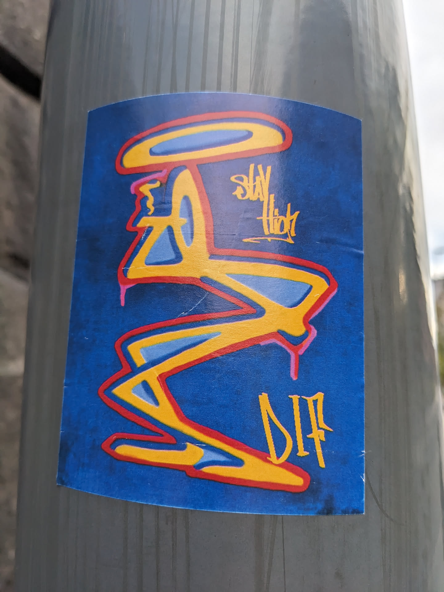 A sticker with an abstract design in yellow, red, and blue on a dark blue background. The text "Stay High" and a tag "DIP" are visible. 