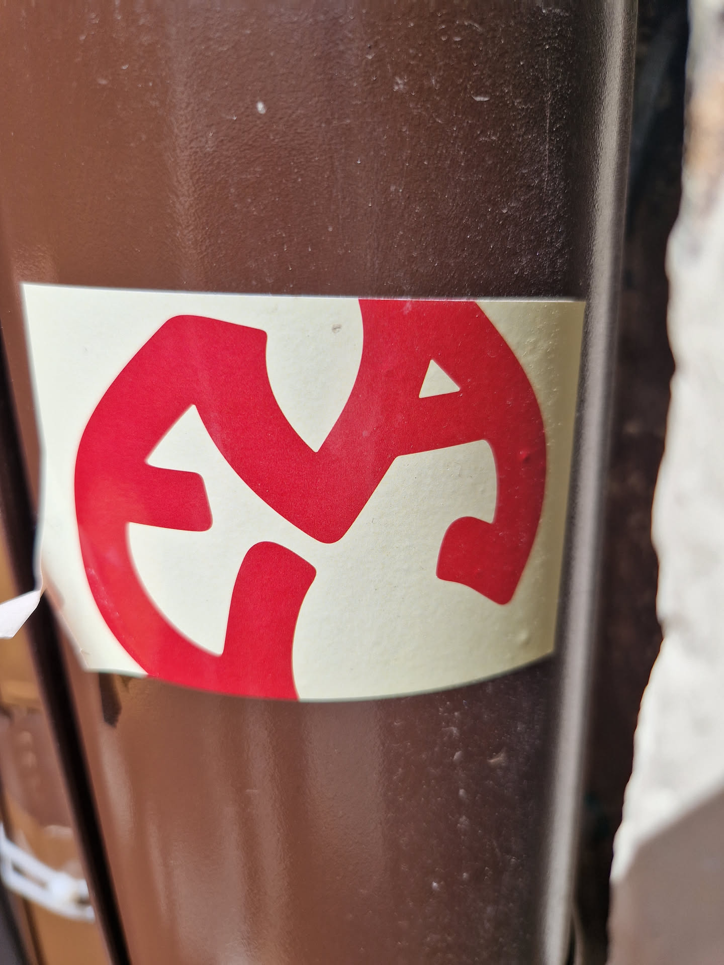 A round sticker with an abstract red symbol on a beige background. The symbol is a stylized design that is not easily recognizable as representing any specific object or concept. The sticker is affixed to a brown cylindrical surface. 