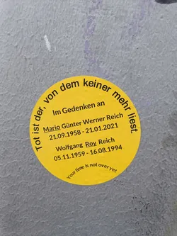 A yellow round sticker with a memorial message for Mario Günter Werner Reich and Wolfgang Roy Reich.  It includes their birth and death dates and the phrase "Your time is not over yet".