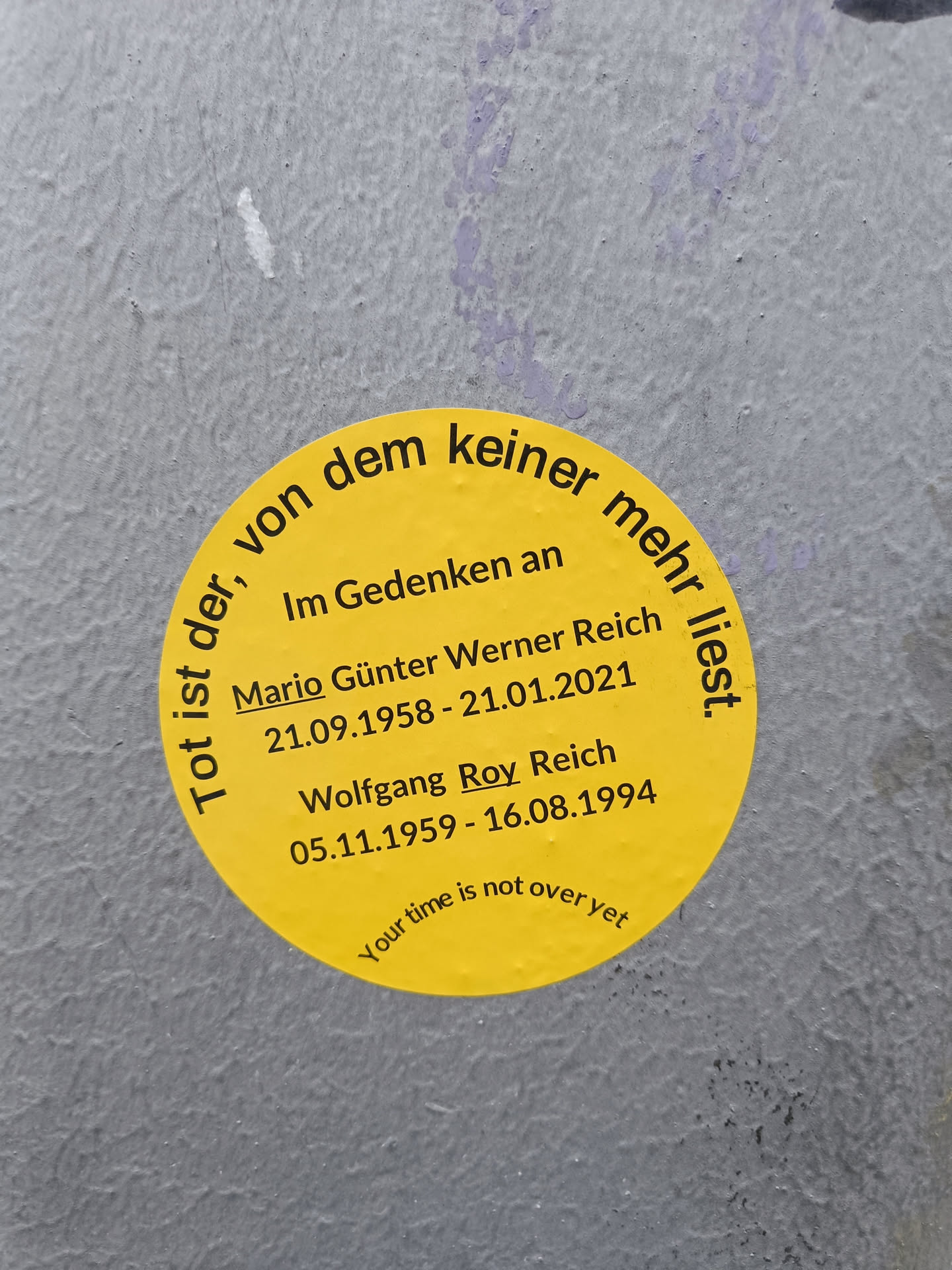 A yellow round sticker with a memorial message for Mario Günter Werner Reich and Wolfgang Roy Reich.  It includes their birth and death dates and the phrase "Your time is not over yet".