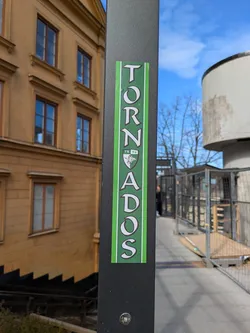 A tall, rectangular sticker with a green background and white text that reads 'TORNADOS' vertically is adhered to a dark gray metal pole. The sticker features a logo in the center, and the overall design is clean and simple. The pole is positioned outdoors, with a yellow building and a blue sky visible in the background.