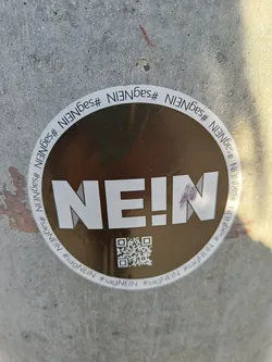 Round gold sticker with the word NEIN (German for NO) in bold white letters.  The sticker also includes a QR code and the hashtag #sagNEIN which means 'say no' in German. The hashtag is repeated around the circumference of the sticker.