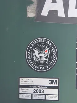 A black and white round sticker with the text "Andreas Leone" around a central emblem. The emblem depicts an eagle and other symbolic elements. The sticker appears to be affixed to a dark green surface, possibly a container or sign.  There is a 3M label nearby, indicating the material of the surface.