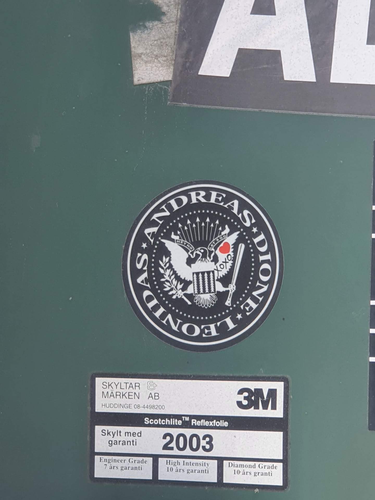 A black and white round sticker with the text "Andreas Leone" around a central emblem. The emblem depicts an eagle and other symbolic elements. The sticker appears to be affixed to a dark green surface, possibly a container or sign.  There is a 3M label nearby, indicating the material of the surface.