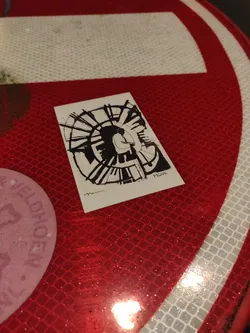 A black and white sticker featuring an abstract drawing of a spiral with a stylized human figure in the center. The style is minimalist, using primarily lines and shading. The sticker appears to be hand-drawn or digitally created to emulate that style. The sticker is affixed to a red reflective surface, possibly a traffic sign.