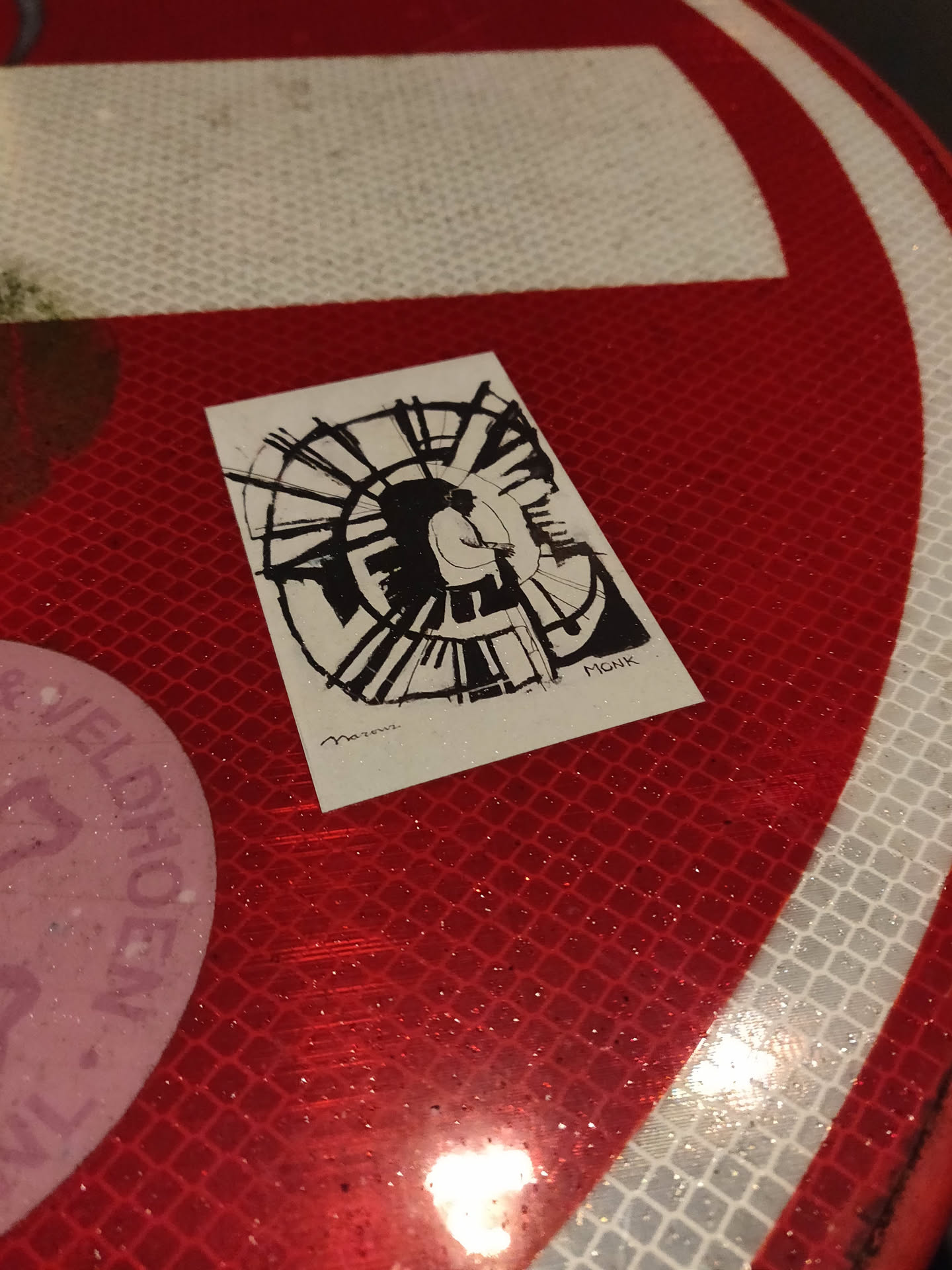 A black and white sticker featuring an abstract drawing of a spiral with a stylized human figure in the center. The style is minimalist, using primarily lines and shading. The sticker appears to be hand-drawn or digitally created to emulate that style. The sticker is affixed to a red reflective surface, possibly a traffic sign.