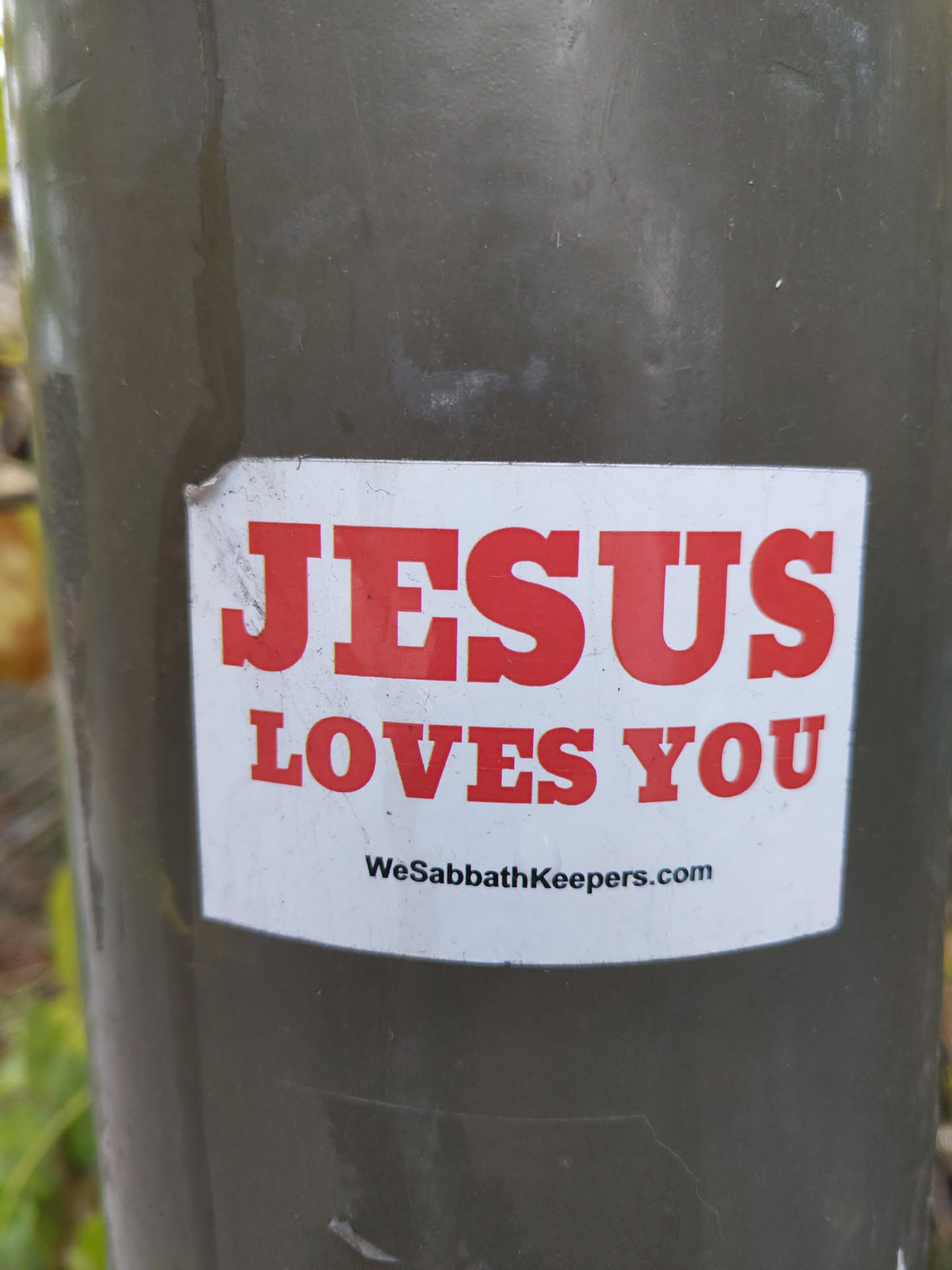A religious sticker with the message "Jesus Loves You" and a website address WeSabbathKeepers.com. The sticker is white with red lettering and is affixed to a dark green pole.