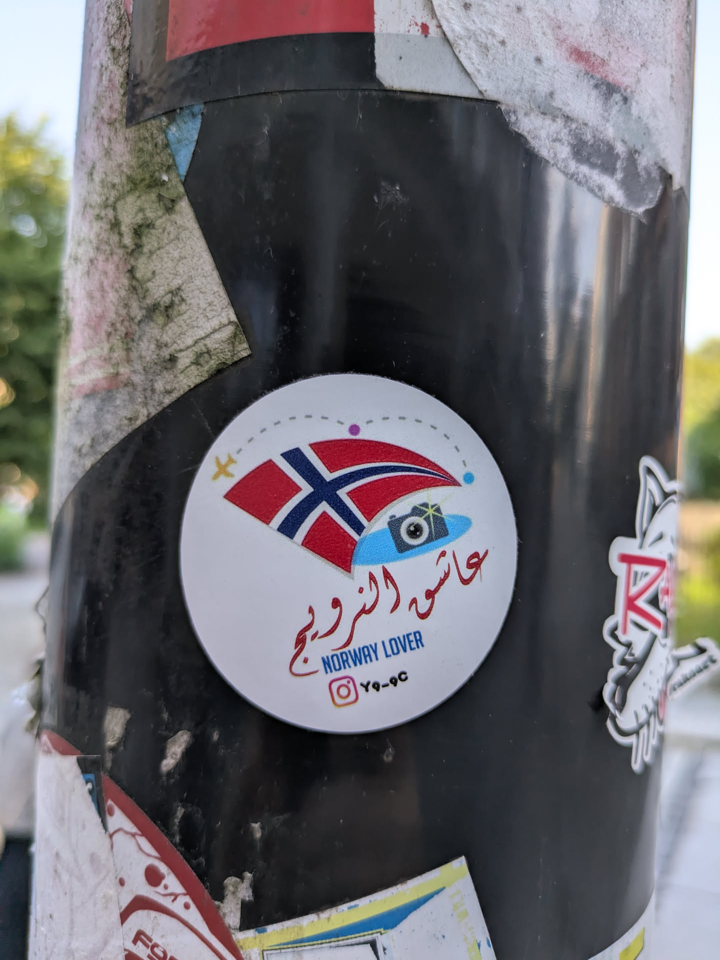 This is a round white sticker affixed to a dark pole. The central design includes a stylized Norwegian flag, appearing to flutter, positioned above a light blue shape that resembles either a cloud or water, featuring a camera icon with a lens flare. Above the flag, a dotted line illustrates an airplane's flight path, complete with a small yellow airplane symbol, suggesting travel. The sticker has text in red Arabic script, "عاشق النرويج" (Aashiq an-Narweej), which translates to "Norway Lover," and below it, "NORWAY LOVER" in blue English text. At the very bottom, there is an Instagram logo followed by the handle "Y9-9C."