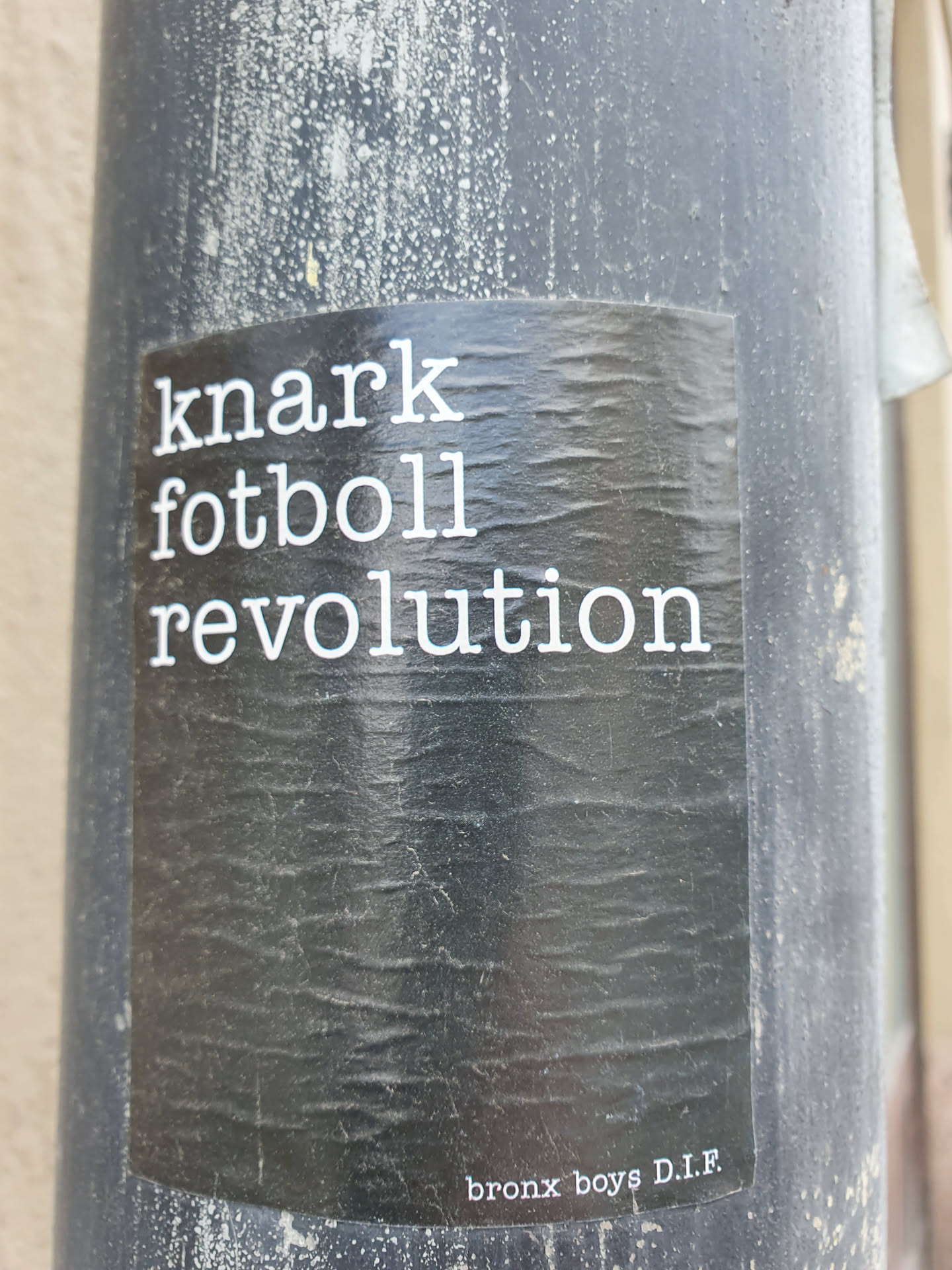 A black and white sticker with the text "knark fotboll revolution" and the words "bronx boys D.I.F." at the bottom. The sticker is affixed to a gray pole and shows some wear.