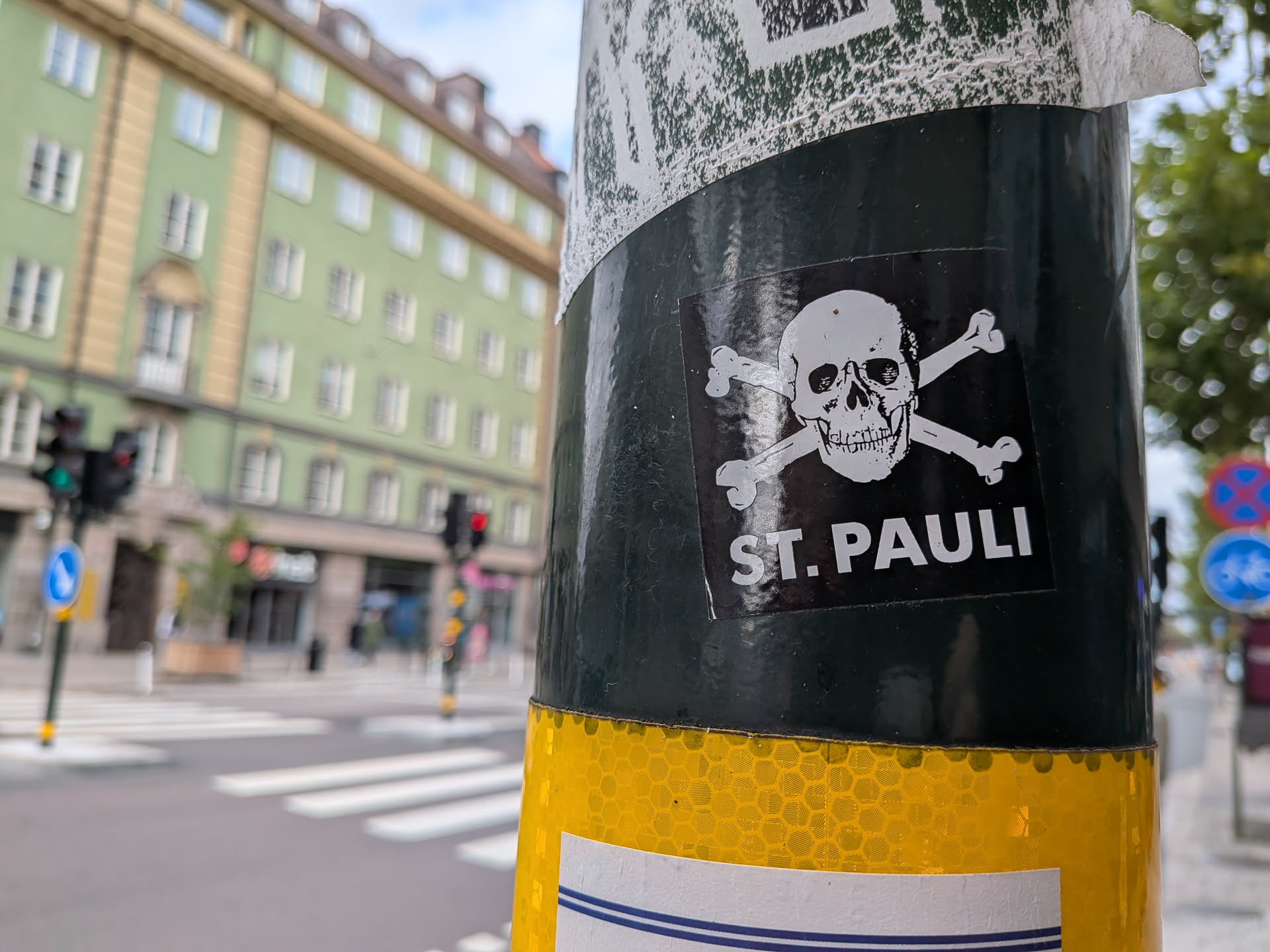 A square black sticker featuring a white skull and crossbones illustration, a symbol associated with the German football club FC St. Pauli. Below the skull, the text 'ST. PAULI' is printed in bold, white, sans-serif capital letters. The sticker is affixed to a dark green metal pole with a yellow reflective band visible below it. The background shows a blurred city street with a green building and a pedestrian crossing.