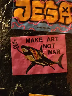 Pink square sticker featuring a stylized shark holding an olive branch in its mouth. The text "MAKE ART NOT WAR" is prominently displayed. The shark is depicted in colorful, striped patterns.